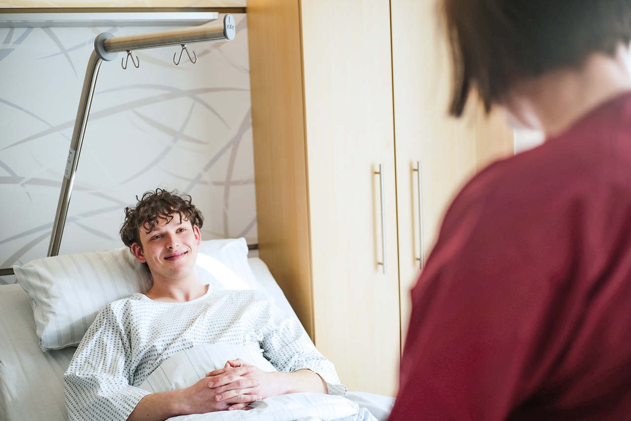 Ein junger Patient im Bett liegend spricht mit einer Pflegerin.