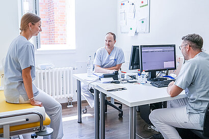 Zwei Ärzte und eine Ärztin in einem Behandlungsraum mit PCs auf dem Tisch.